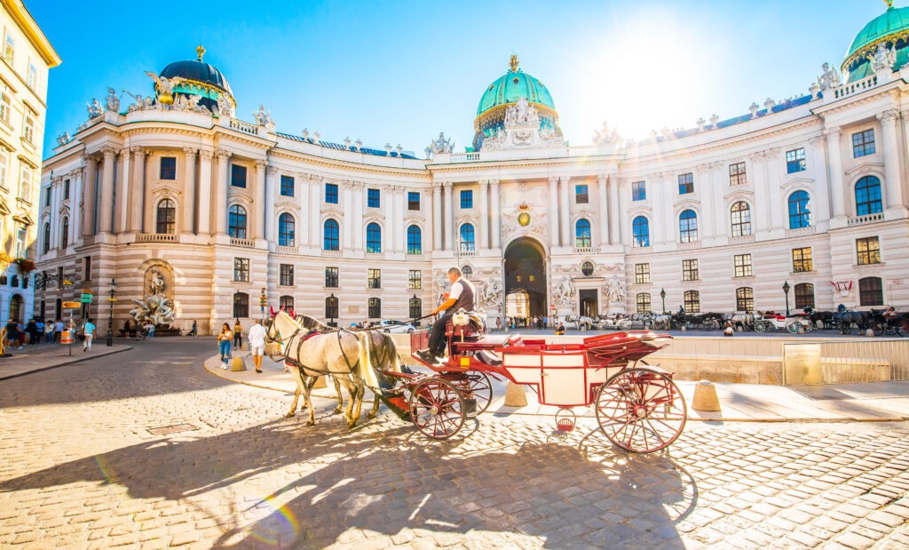 Die Hofburg in Wien mit prächtiger historischer Fassade, davor eine traditionelle Pferdekutsche auf einer sonnigen Straße.