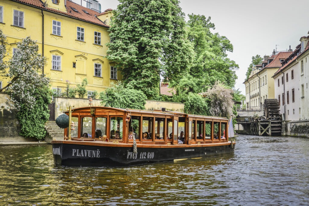 Die Insel Kampa mit dem Fluss Čertovka, umgeben von historischen Gebäuden und einem Oldtimer-Ausflugsboot, das malerisch durch den ruhigen Wasserkanal fährt.