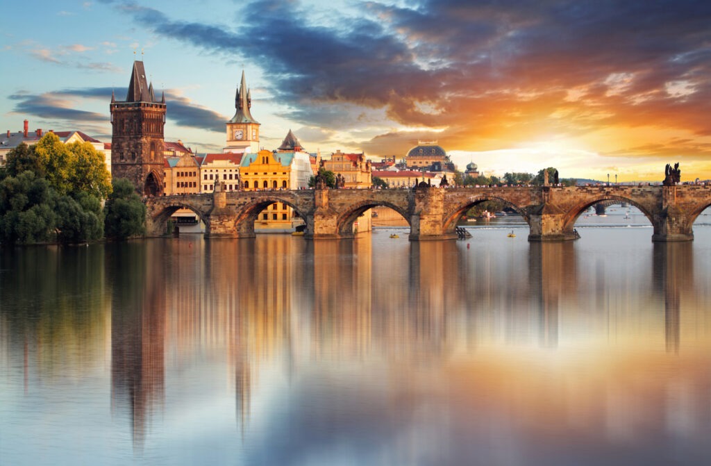 Die Karlsbrücke in Prag, Tschechien, bei Sonnenuntergang, mit ihren historischen Statuen und Blick auf die Prager Burg im Hintergrund, während sich das warme Licht im Wasser der Moldau spiegelt.