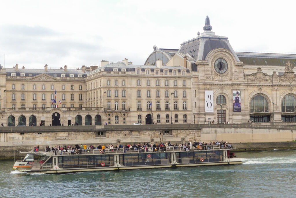 Das Musée d'Orsay am Ufer der Seine, ein beeindruckendes Gebäude im Beaux-Arts-Stil mit markanter Uhr und reflektierenden Wasserflächen.