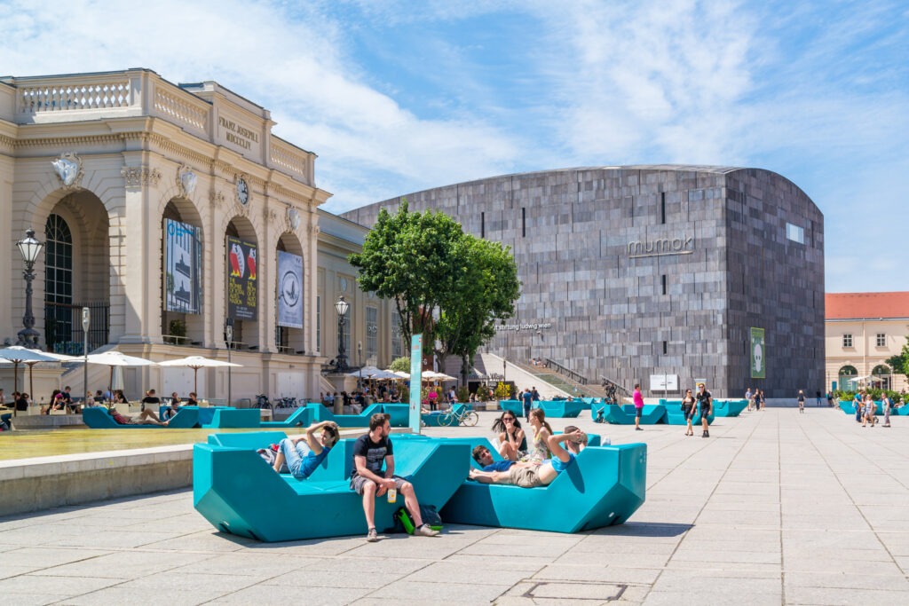 Das MuseumsQuartier in Wien mit moderner Architektur, belebtem Platz und Menschen, die auf den ikonischen Sitzmöbeln entspannen.