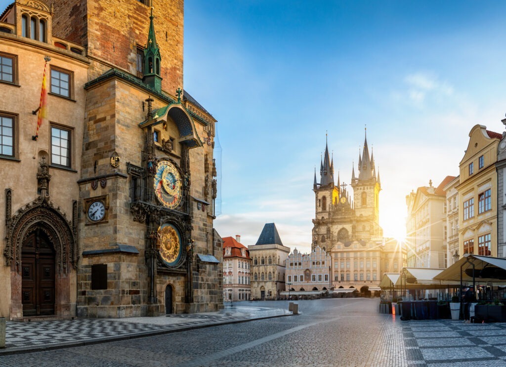 Blick auf die Astronomische Uhr am Prager Rathaus und den Altstädter Ring bei Sonnenaufgang, mit historischem Kopfsteinpflaster und bunten Gebäuden.