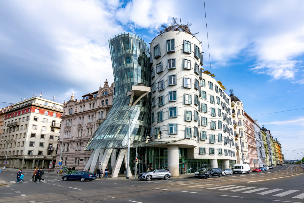 Das berühmte Tanzende Haus in Prag, Tschechien, mit seiner modernen, wellenförmigen Architektur, umgeben von historischen Gebäuden, bei blauem Himmel und Sonnenschein.