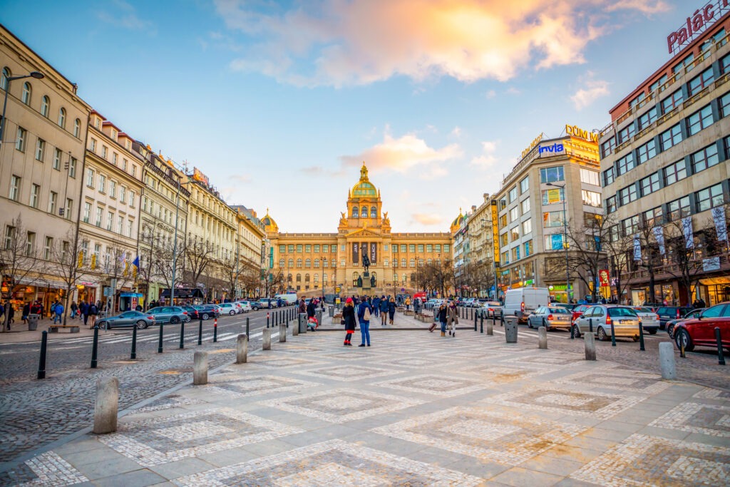 Menschen flanieren über den Wenzelsplatz bei Sonnenuntergang, während die Straßenlaternen und historischen Gebäude in warmem Licht erstrahlen.