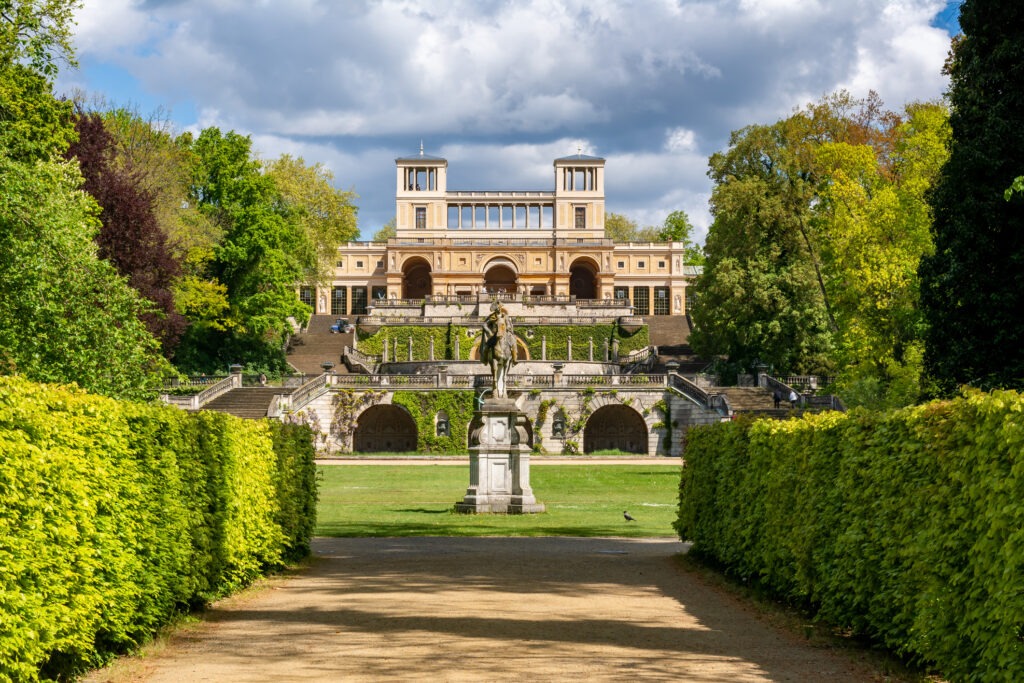 Friedrich-der-Große-Denkmal im Park Sanssouci, Potsdam, mit der Orangerie im Hintergrund und umgeben von historischer Gartenlandschaft.