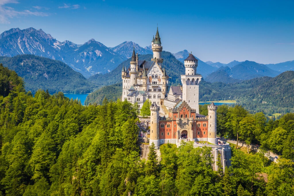 Schloss Neuschwanstein in Bayern, umgeben von malerischer Berglandschaft und herbstlich gefärbten Wäldern bei strahlendem Sonnenschein.